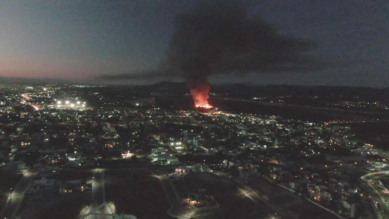 Aerial frame over San Jose Del Cabo at nightfall with dense city lighting, a bright red fire in the background, a broad black smoke plume, and no visible people.