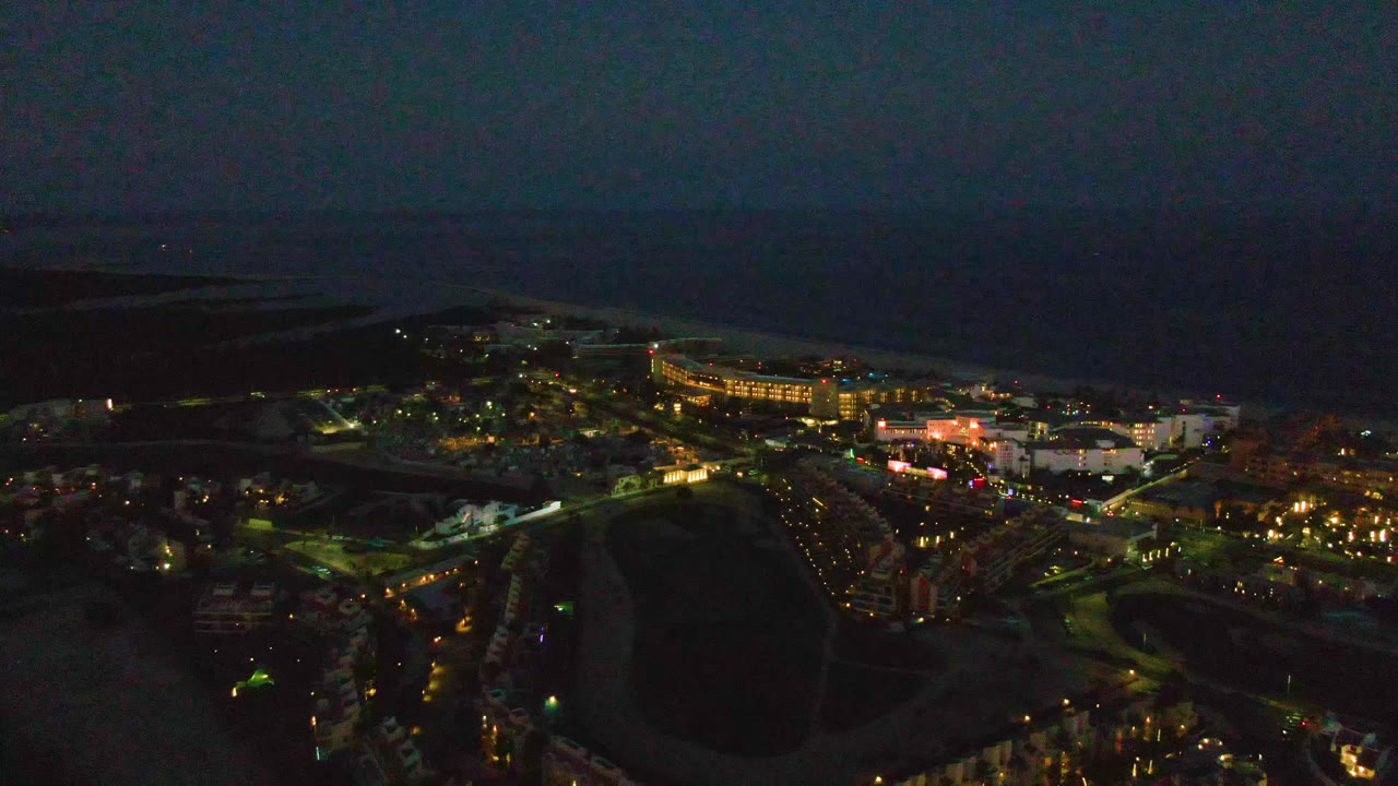 Opening night aerial over San Jose del Cabo with warm town lights, streets, buildings, a dark beach, and ocean beyond, with no visible people.