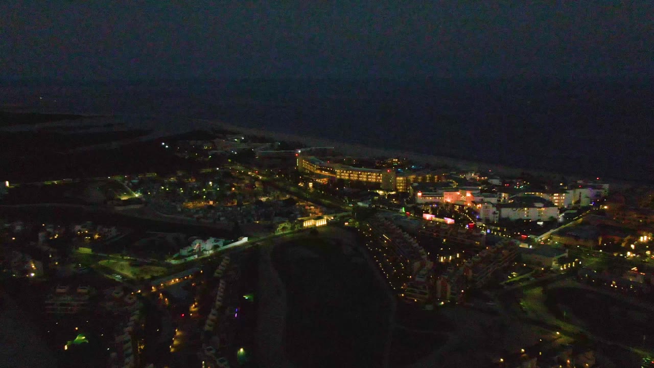 Mid-clip aerial over San Jose del Cabo with scattered warm lights across buildings and roads, a black ocean horizon, and no visible people.