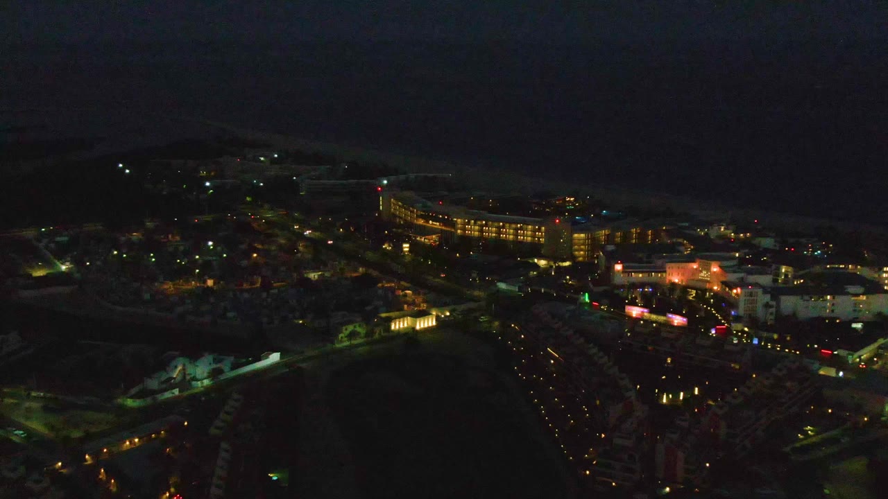 Night aerial view over San Jose del Cabo with scattered city lights, a curved lit building at right, dark ocean beyond, and no visible people.