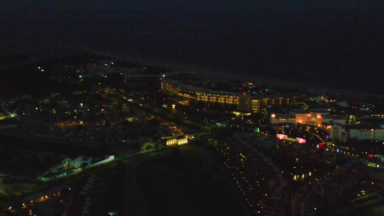 Aerial nighttime frame of San Jose del Cabo showing warm-lit buildings, green street lighting, traffic traces on roads, dark water in the background, and no visible people.