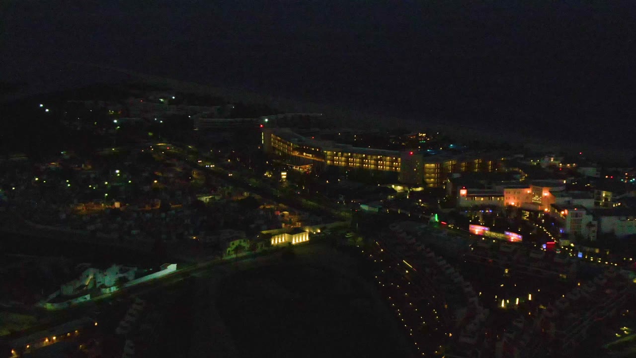 San Jose del Cabo appears from above at night with a bright curved building on the right, dense streetlights through the center, dark shoreline beyond, and no visible people.