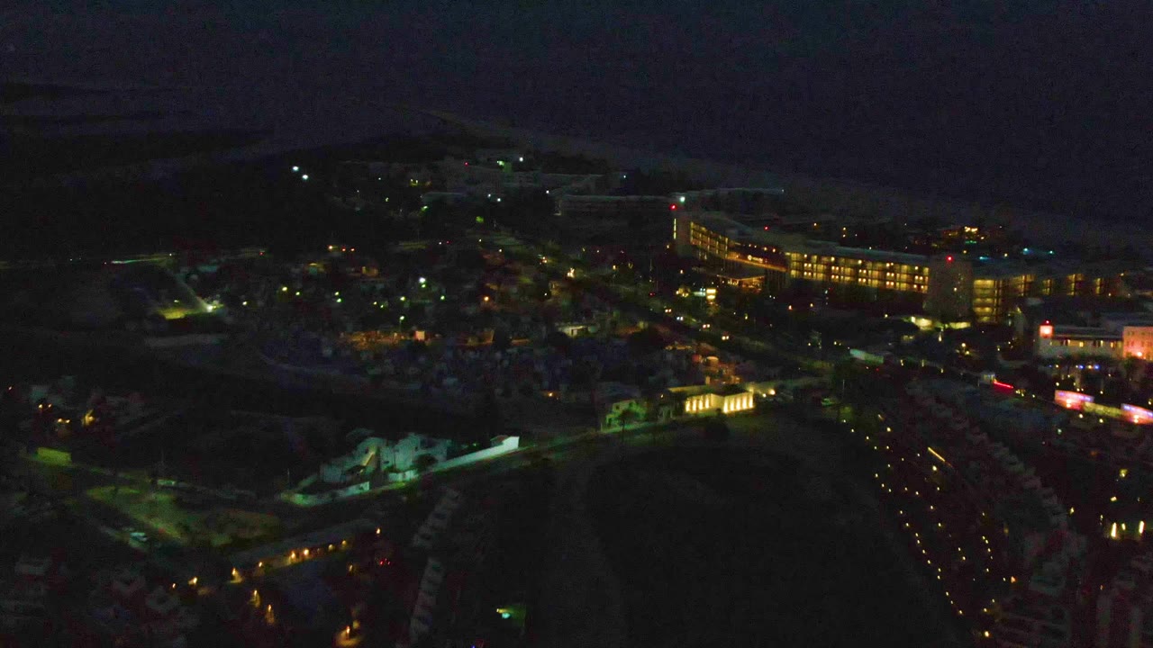 San Jose Del Cabo aerial view one second in, with lit road lines, clustered buildings, dark foreground terrain, ocean in the distance, and no visible people.