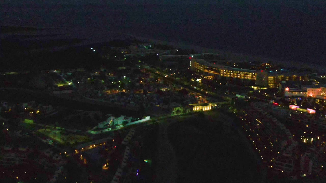 San Jose Del Cabo from an elevated drone-like angle, showing bright building windows, road lights, darker beachside ground, ocean horizon, and no visible people.