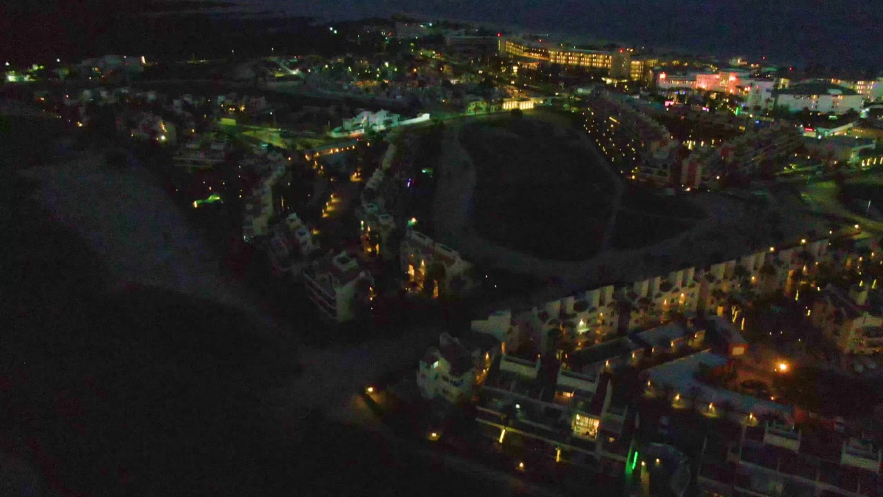 Early frame of San Jose Del Cabo at night from above, with a dark shoreline at left, lit buildings below, distant town lights, and no visible people.