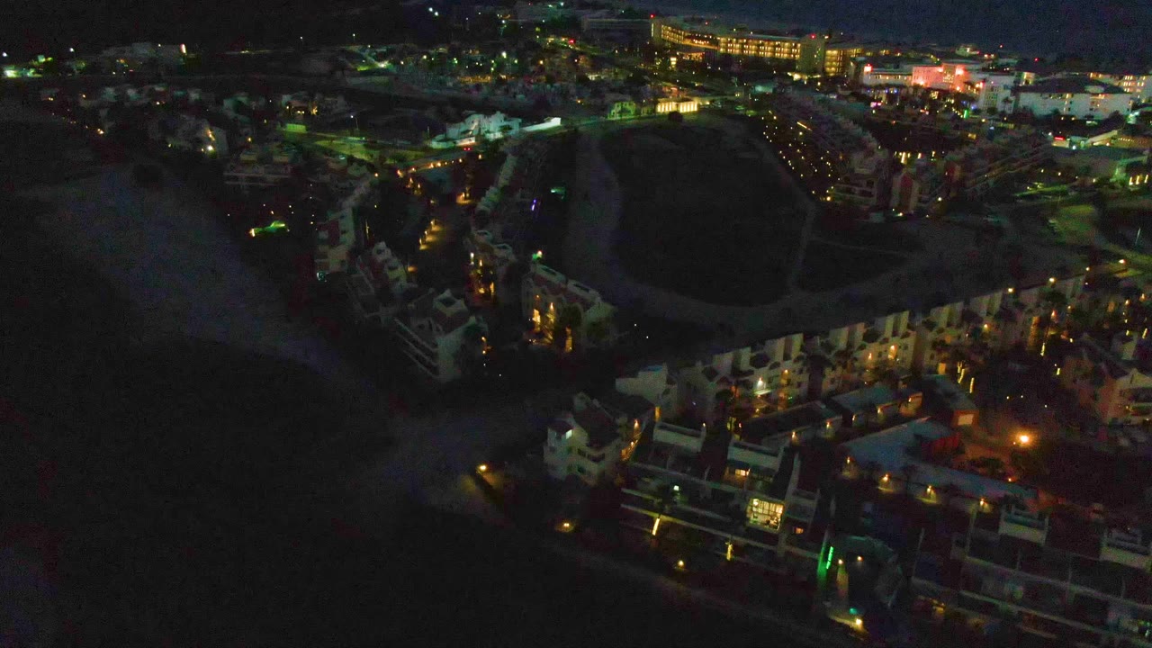 San Jose Del Cabo aerial view one second in, showing the beach and water in shadow, bright street lines, lit waterfront buildings, and no visible people.