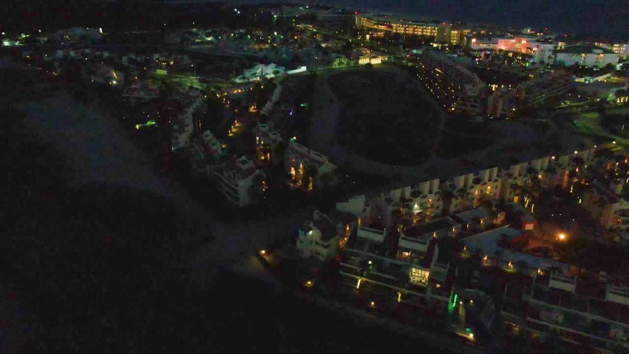 Mid-early frame over San Jose Del Cabo with waterfront buildings in the lower frame, a dark beach beside them, scattered city lights beyond, and no visible people.