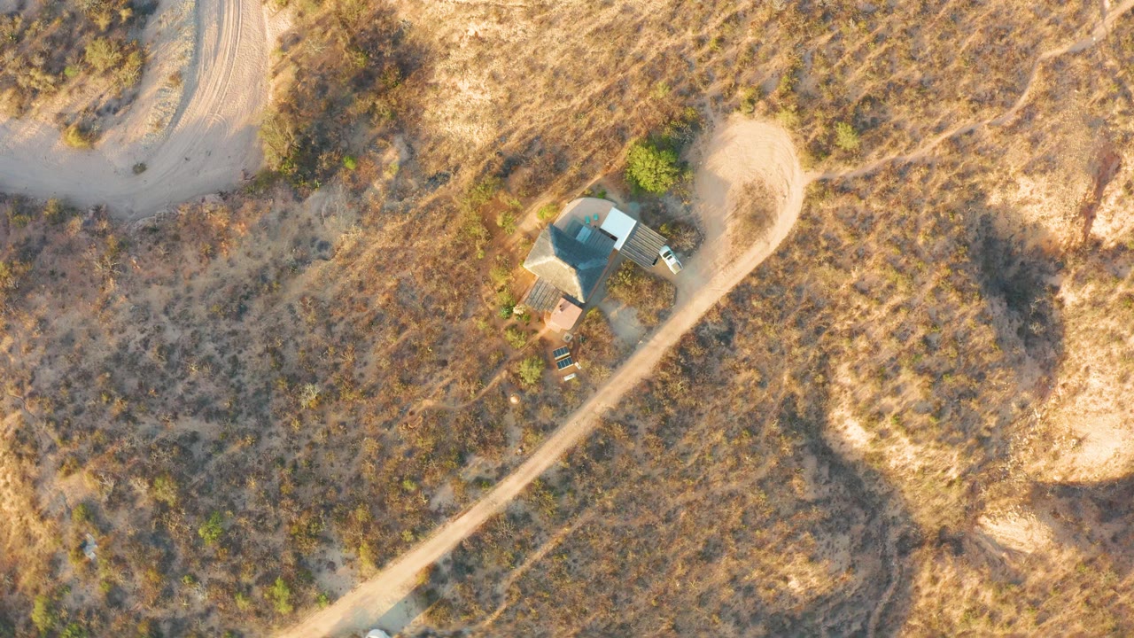 Overhead drone frame in Cabo Pulmo, Baja California Sur, showing a small car entering a pale dirt road near dry grass, shrubs, and a blue-roofed structure, with no visible people.