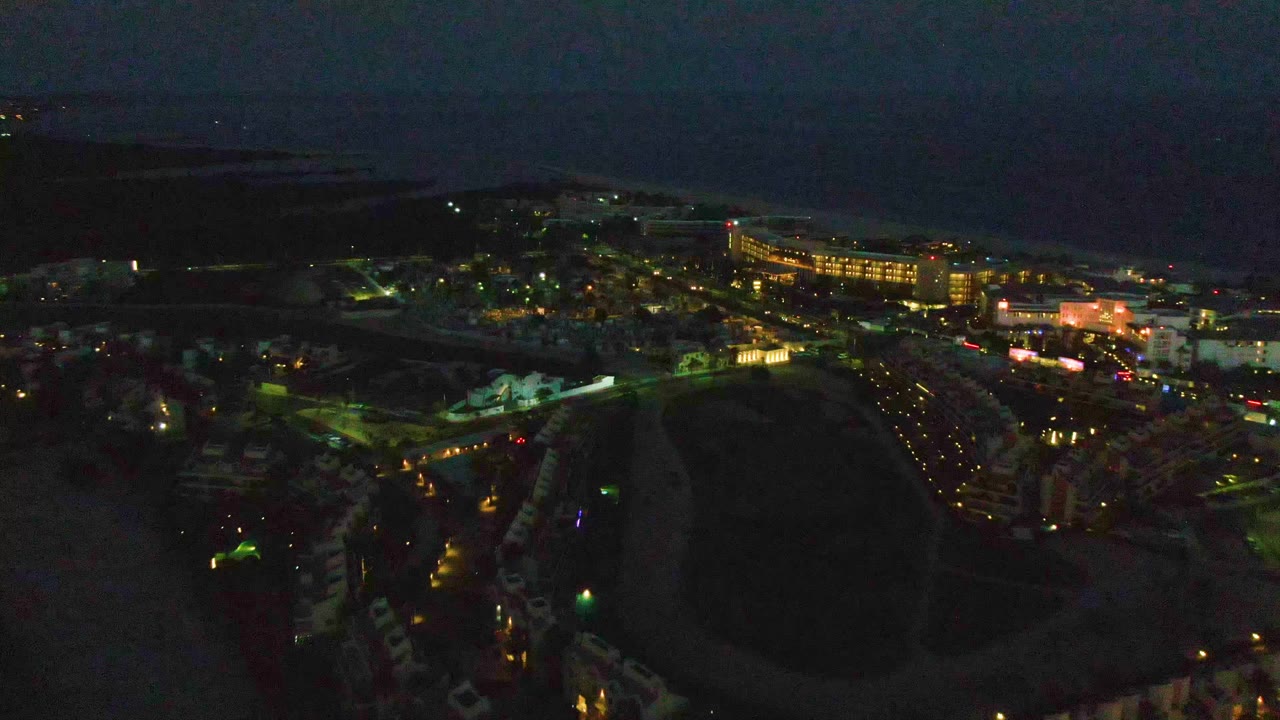 Night aerial frame over San Jose del Cabo with a dark open field below, lit streets and buildings beyond, ocean on the horizon, and no visible people.
