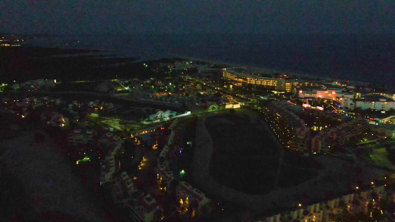 Aerial nighttime view in San Jose del Cabo with foreground rooftops, a large unlit field, brighter street grids, ocean beyond town, and no visible people.
