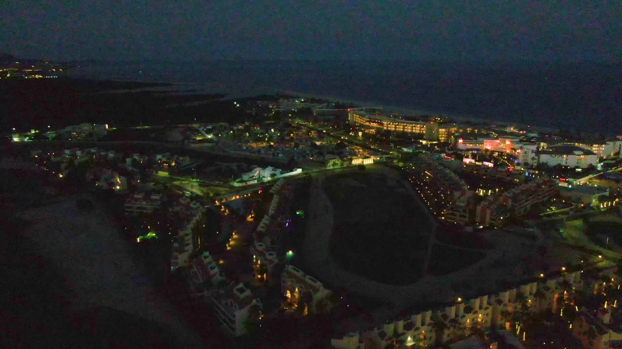 Night drone-style view over San Jose del Cabo showing a broad dark field in front, illuminated town blocks, a brighter commercial strip, ocean in the distance, and no visible people.