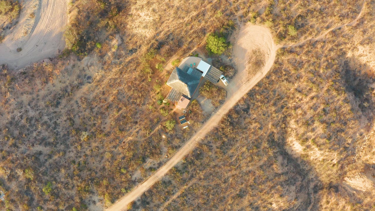 Overhead drone view in Cabo Pulmo, Baja California Sur, showing a small roofed structure beside a pale dirt road amid dry grass and scattered bushes, with no visible people.