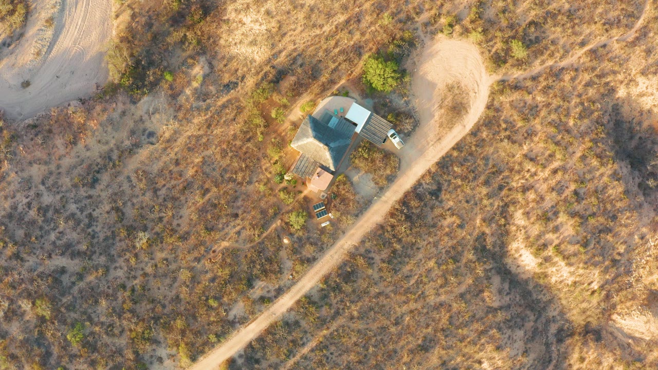 Top-down view in Cabo Pulmo, Baja California Sur, showing dry tan grassland, scrub, a small roofed structure, and the dirt road cutting diagonally through the frame, with no visible people.