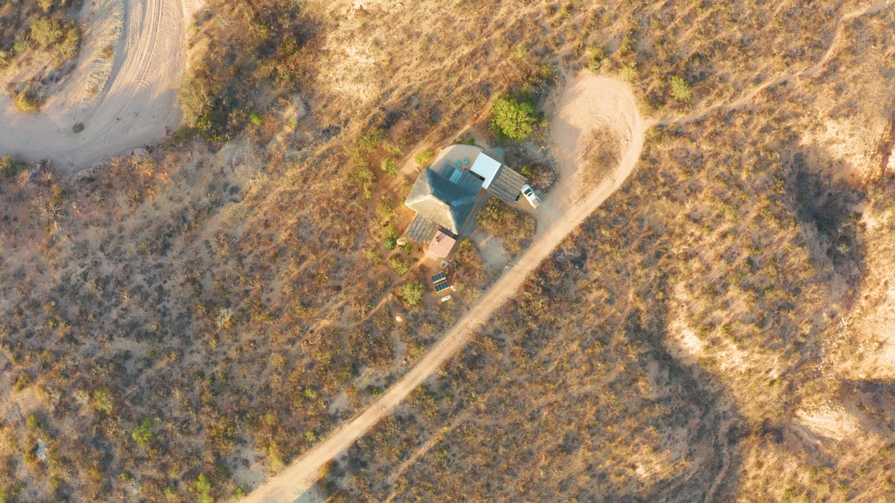 Drone view over Cabo Pulmo, Baja California Sur, showing a vehicle beside a small roofed structure and a curving dirt road through dry grass, with no visible people.