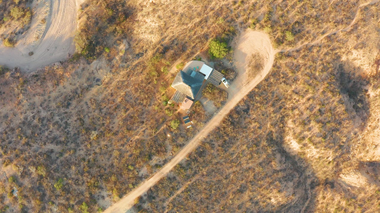 Aerial view of a dirt road, dry brush, and a small building with a vehicle nearby.