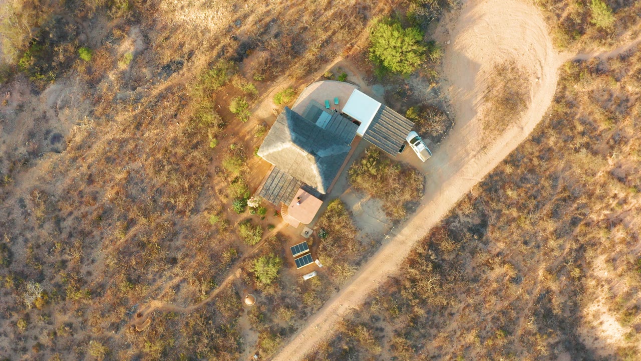 Overhead view in Cabo Pulmo, Baja California Sur, showing a small isolated house, diagonal dirt road, dry grass, scattered green shrubs, solar panels, a white vehicle, and no visible people at the start of the clip.