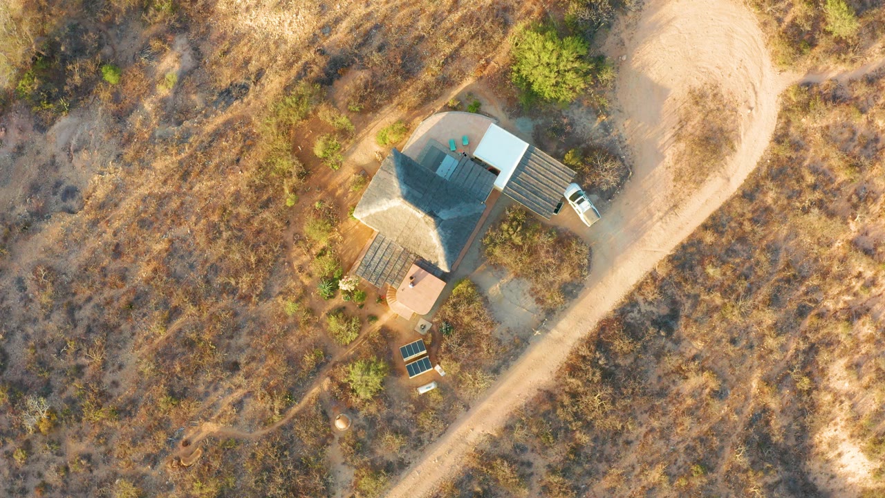 Aerial view of the same rural house with dry grass, shrubs, and a pale dirt road.
