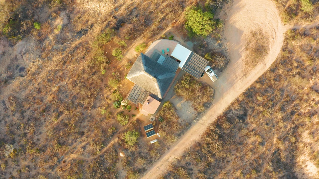At 0:02, the Cabo Pulmo aerial view shows the isolated house beside the curving dirt road, a white vehicle near the roofline, solar panels below the structure, warm light, and no visible people.