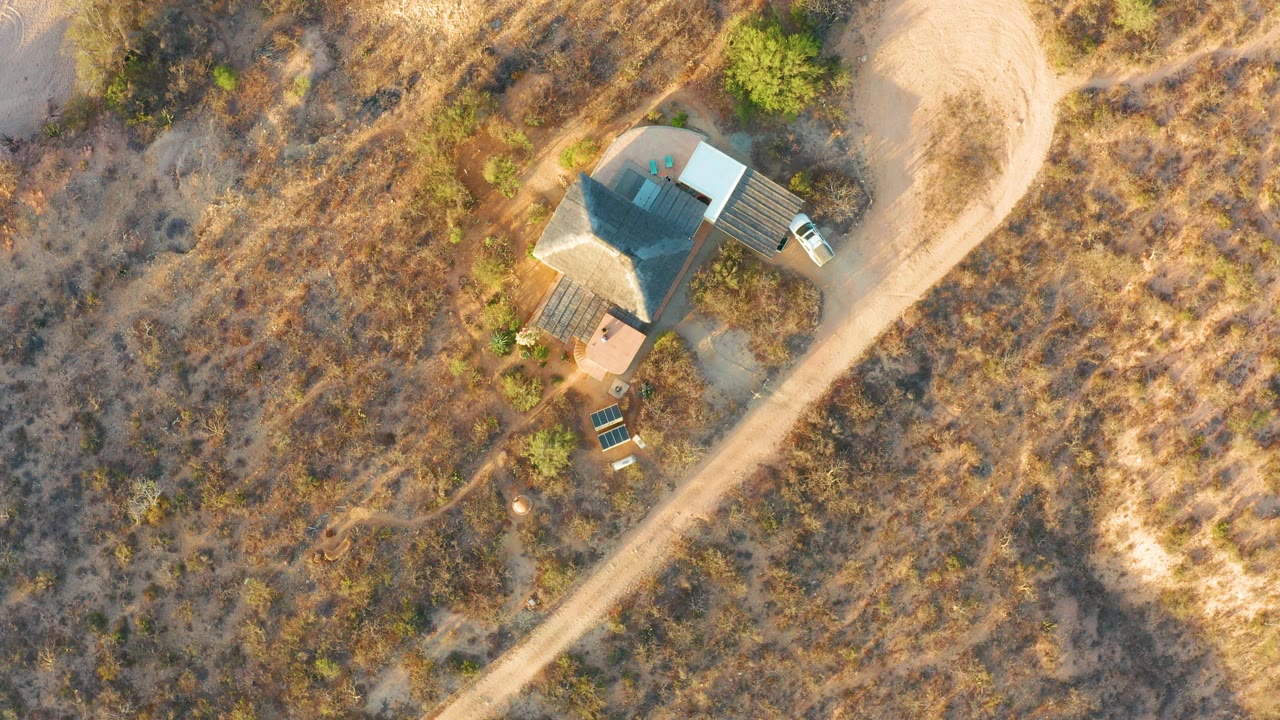 Overhead frame in Cabo Pulmo, Baja California Sur showing a small house with a dark roof, a pale dirt road, parked vehicles, dry vegetation, and no visible people.