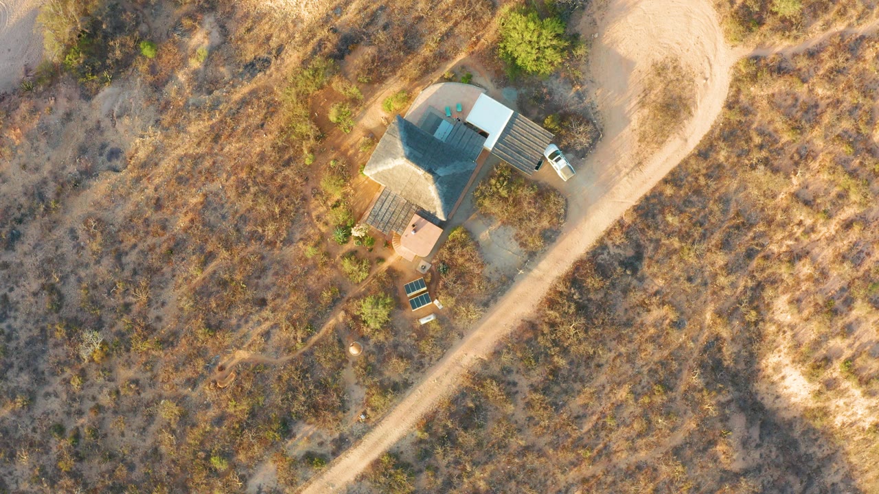 Second overhead frame in Cabo Pulmo shows the house roof still dark, the dirt road curving past the property, two parked vehicles, surrounding scrub, and no visible people.