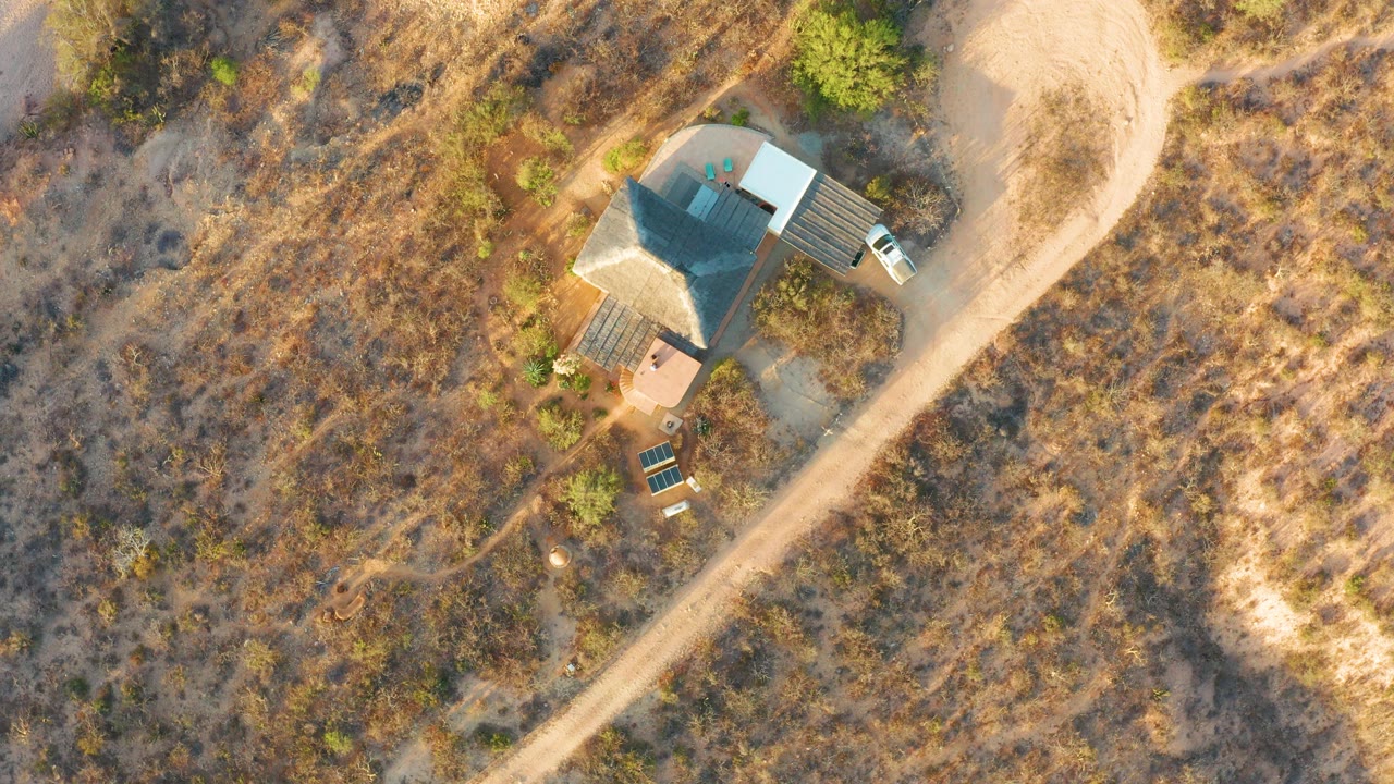 Third overhead frame in Cabo Pulmo shows the standalone house among dry grass, a light dirt road along the right side, parked vehicles by the structure, and no visible people.