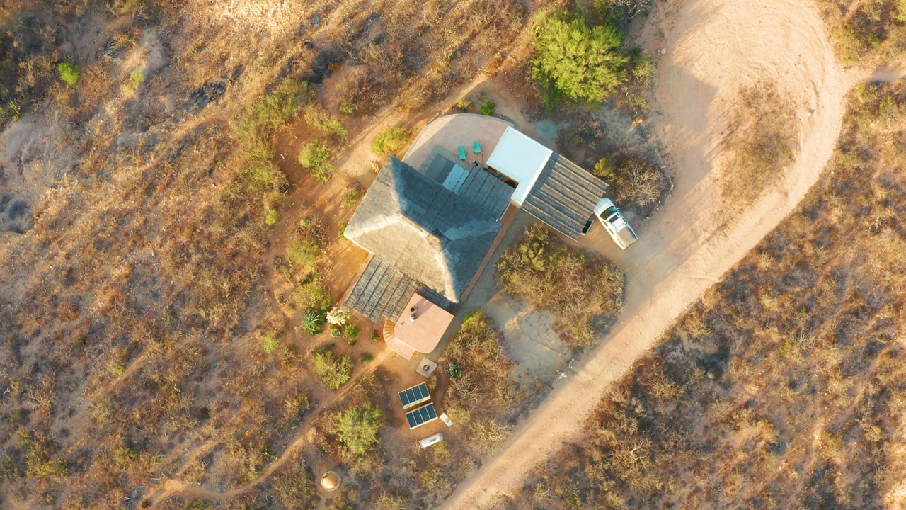 At 0:00, an overhead drone view in Cabo Pulmo shows a small house, a nearby white vehicle, dry brush, scattered trees, and no visible people.