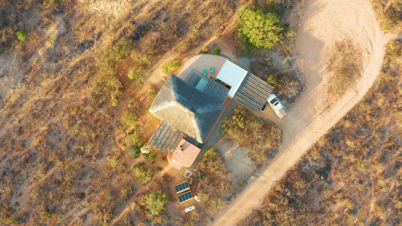 At 0:02, the overhead Cabo Pulmo view centers the roof shadow, deck structures, surrounding dry vegetation, dirt road, and no visible people.