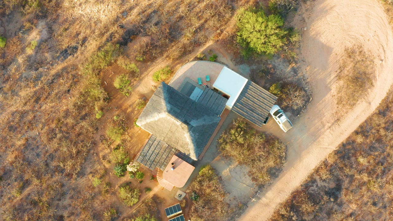 Overhead frame in Cabo Pulmo, Mexico, showing a dark-roofed house beside a pale dirt road, dry vegetation, green shrubs, a white vehicle near the right side, and no visible people.