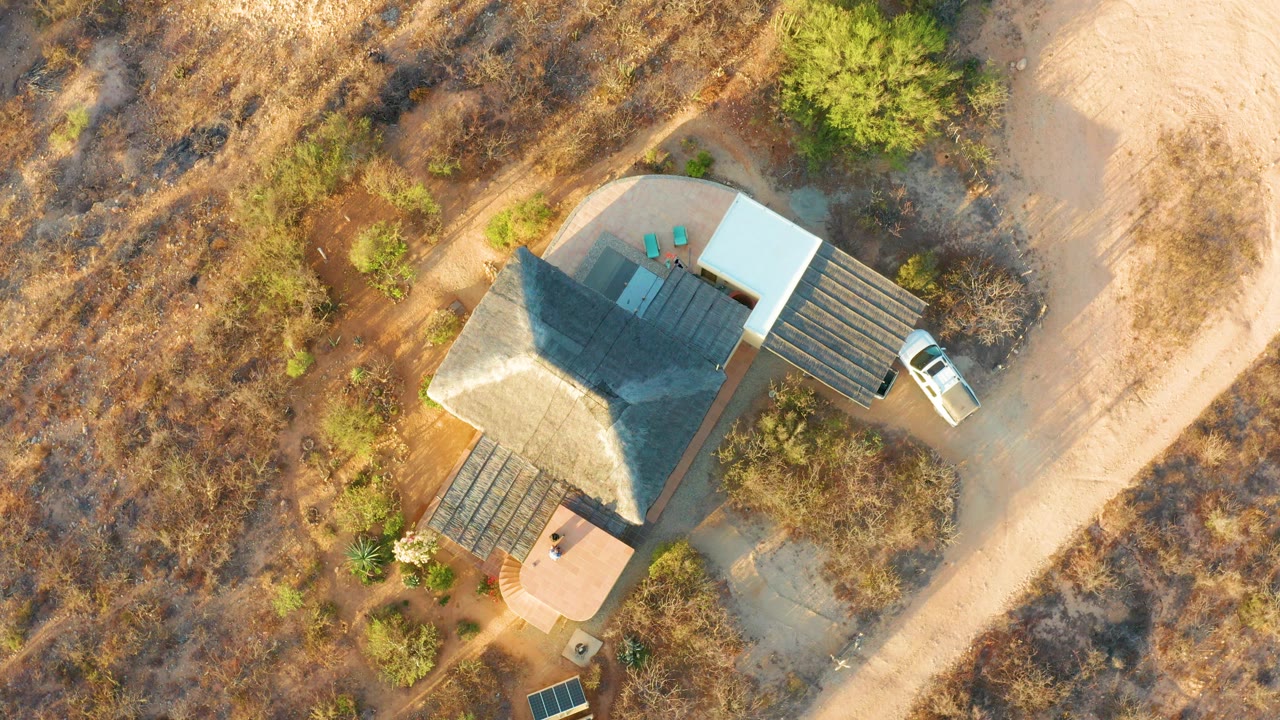 At two seconds, the overhead Cabo Pulmo frame shows the white vehicle slightly farther along the road beside the house, with brown terrain, scattered green shrubs, and no visible people.