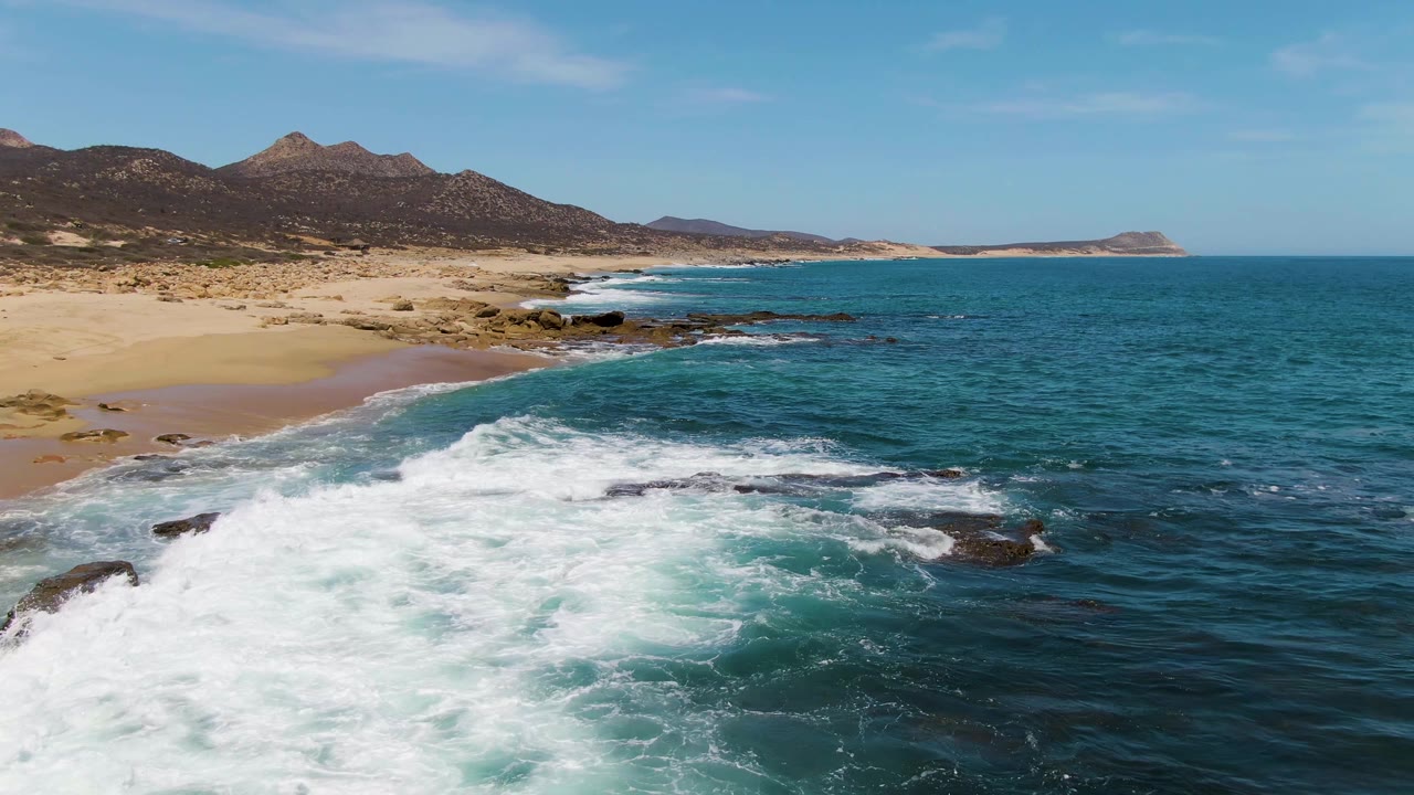 Aerial view of waves breaking over rocks beside a sandy beach and dry hills.