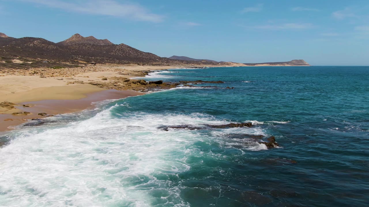 Aerial view at Buzzards Beach showing a broad sheet of white foam crossing the rocky shoreline, sandy beach along the left edge, teal water offshore, low dry hills beyond, and no visible people.