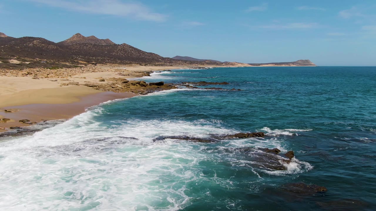 Aerial frame from Buzzards Beach with whitewater covering the center foreground rocks, a curving sandy shoreline to the left, darker blue open water to the right, dry hills behind the beach, and no visible people.