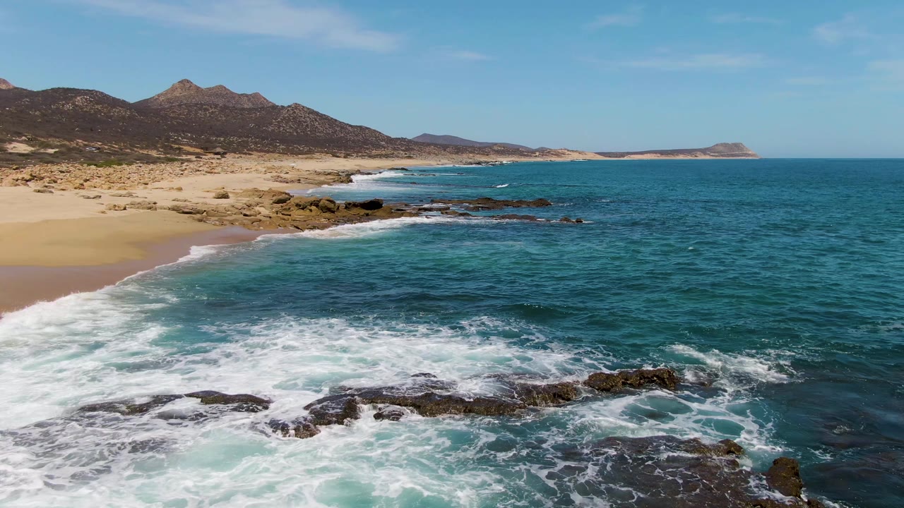 Buzzards Beach aerial frame where the whitewater has moved lower in the foreground, exposing more dark rocks and blue water, with pale sand and dry hills continuing across the background and no visible people.