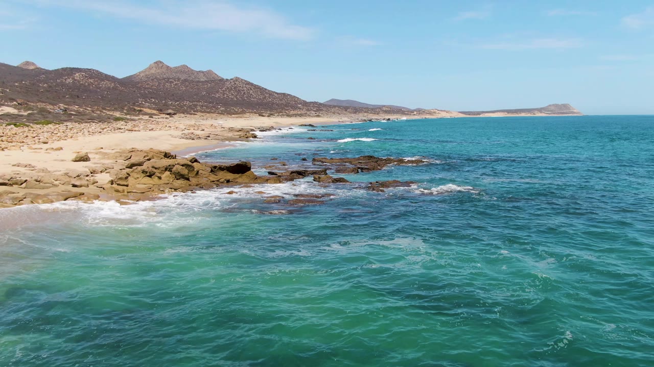 Aerial view at Buzzards Beach, Baja California Sur, with turquoise water in the foreground, white surf near dark rocks, sandy beach at left, mountains in the background, and no visible people.