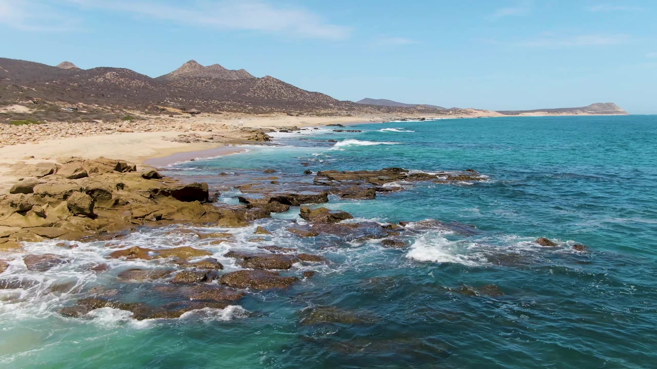 Aerial coastal frame from Buzzards Beach, Baja California Sur, showing more exposed rock shelves in the midground, foamy water around them, sand to the left, distant headland, and no visible people.