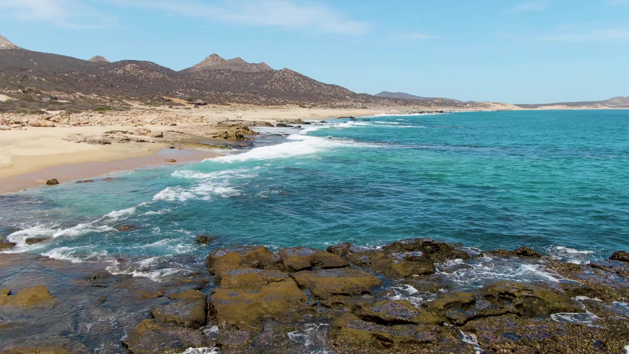 Turquoise waves reaching rocky shore beside a sandy beach and distant mountains.