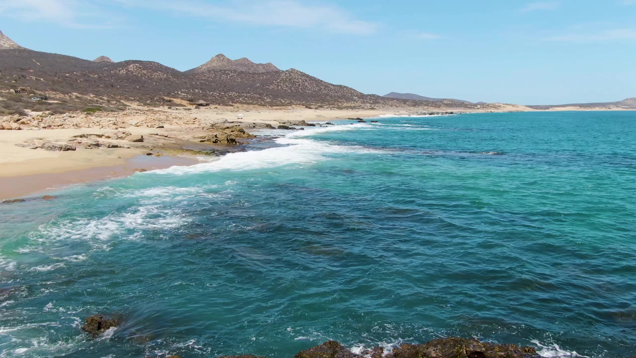 At Buzzards Beach in Baja California Sur, a white wave line runs along the sand as clear turquoise and deeper blue water fills the foreground, with no visible people.