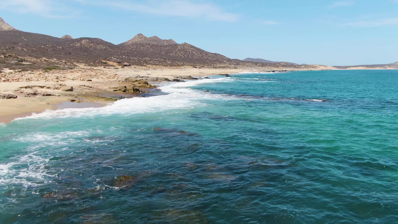 At Buzzards Beach in Baja California Sur, shallow turquoise water passes over dark submerged rocks near the beach, with arid mountains beyond and no visible people.