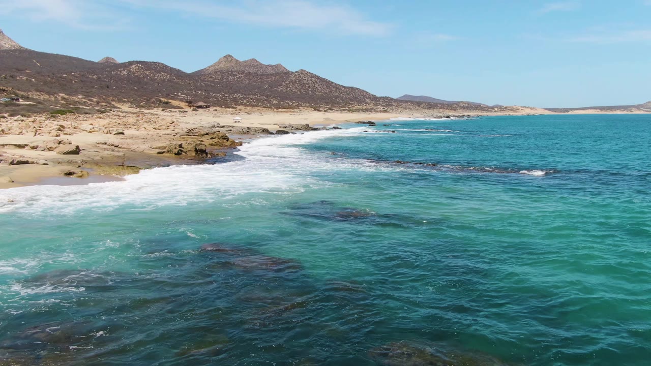 At Buzzards Beach in Baja California Sur, broad white foam pushes across pale turquoise shallows beside the sand, with rocky edges and no visible people.