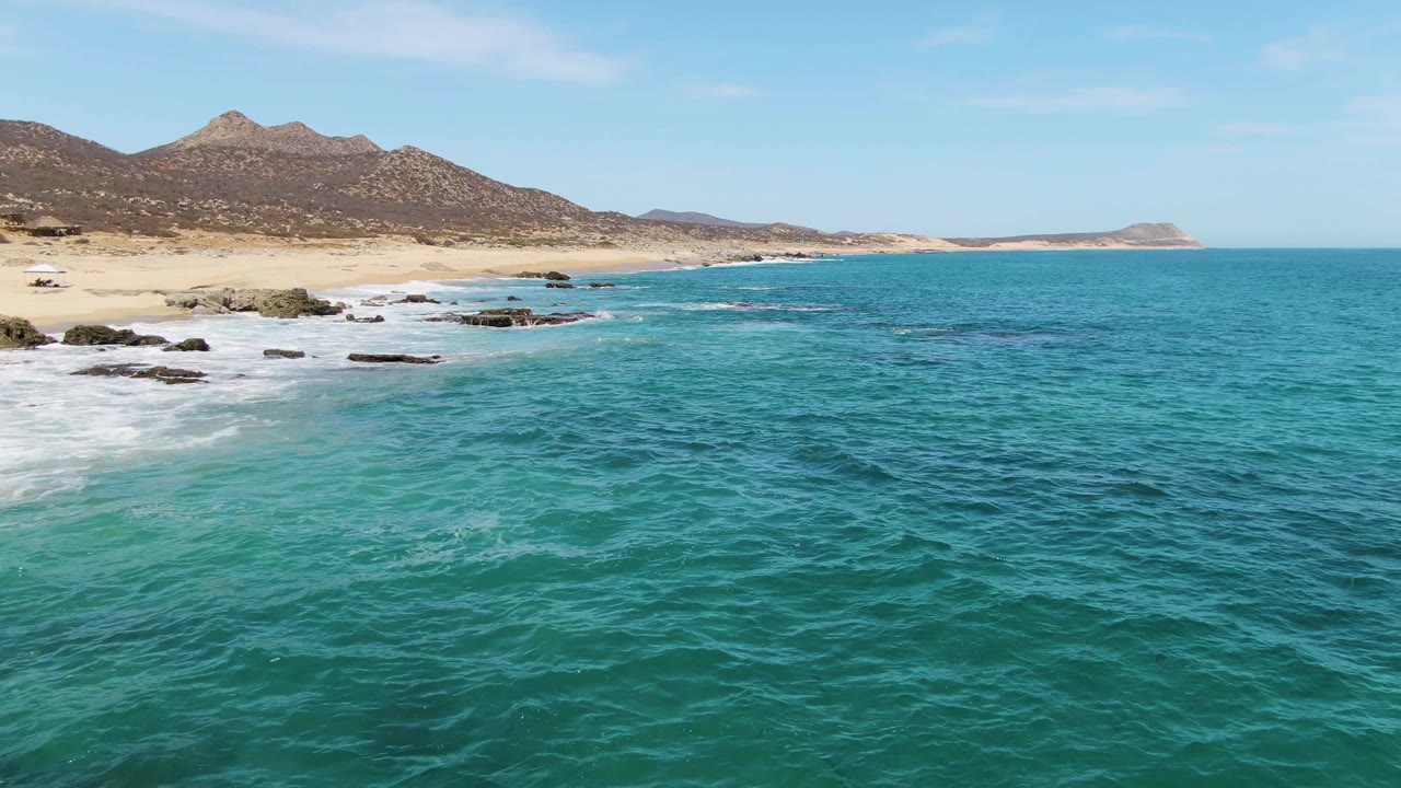 Aerial view over turquoise water at Buzzards Beach, Baja California Sur, with white surf reaching dark rocks, sandy shoreline, brown mountains, clear blue sky, and no visible people.