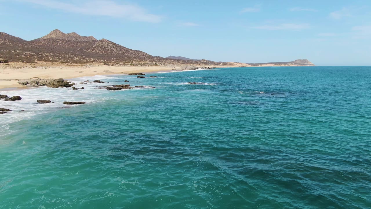 Aerial frame at Buzzards Beach showing more open turquoise water in front, white foam tracing the rocky shoreline, pale sand below the mountains, and no visible people.