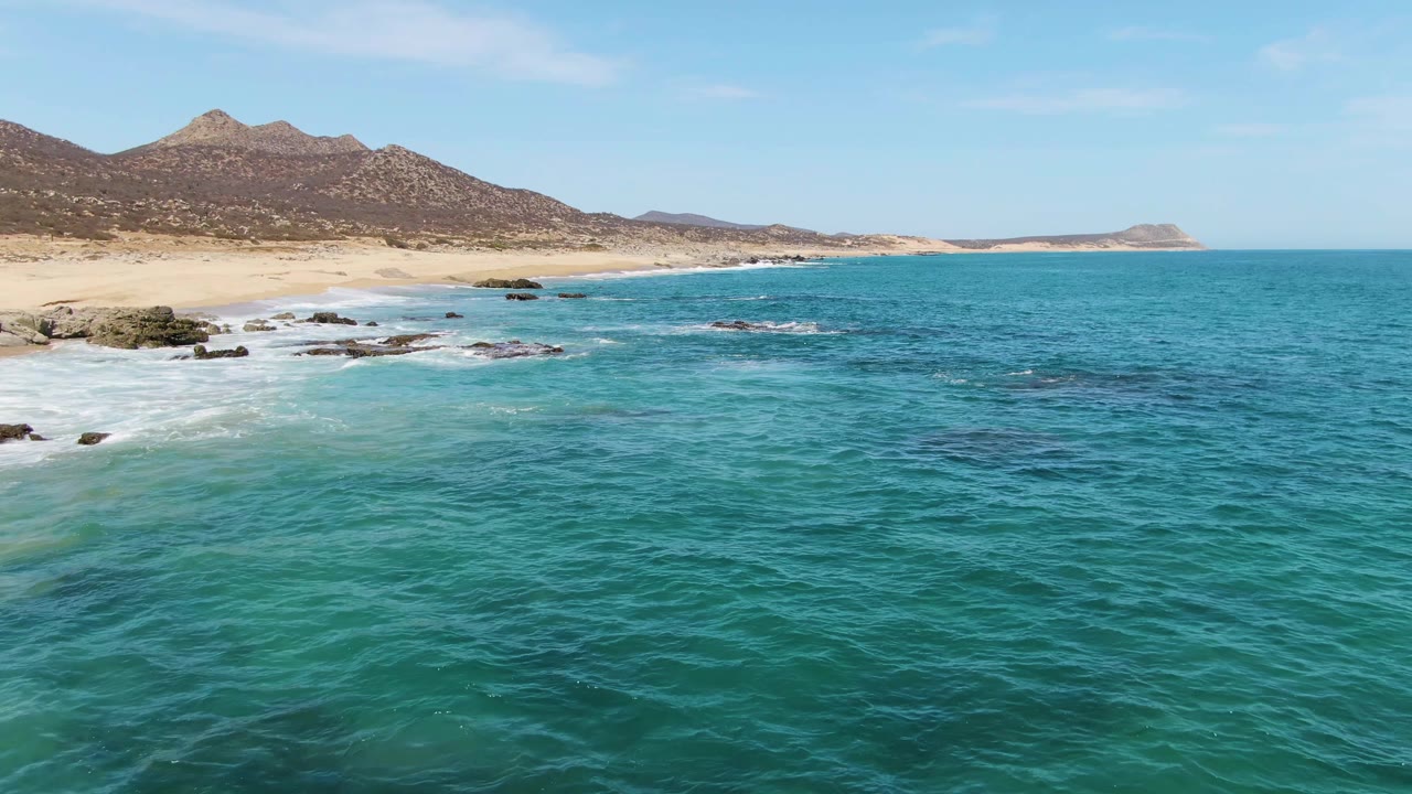 Wide aerial view at Buzzards Beach with textured blue-green ocean across the foreground, small white breaks near rocks at left, sandy beach, mountain ridge, and no visible people.