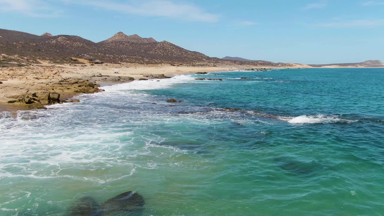 Turquoise waves breaking over rocky shoreline below distant mountains.