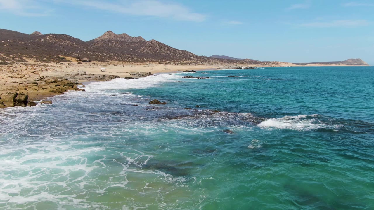 At 0:01, Buzzards Beach surf spreads wider across shallow turquoise water near exposed rocks, with sandy coast and mountains behind and no visible people.