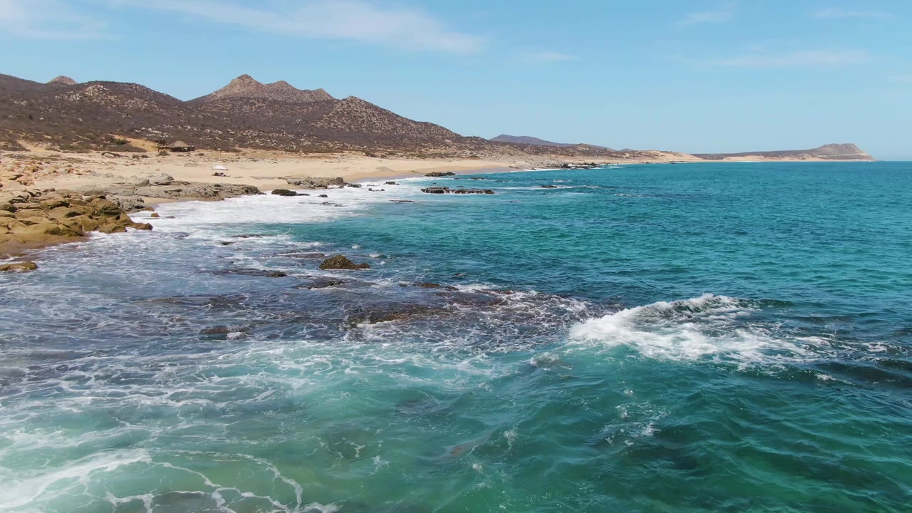 At 0:02, a band of white surf curls over the rocky edge of Buzzards Beach, with deeper blue water to the right, sandy beach beyond, and no visible people.
