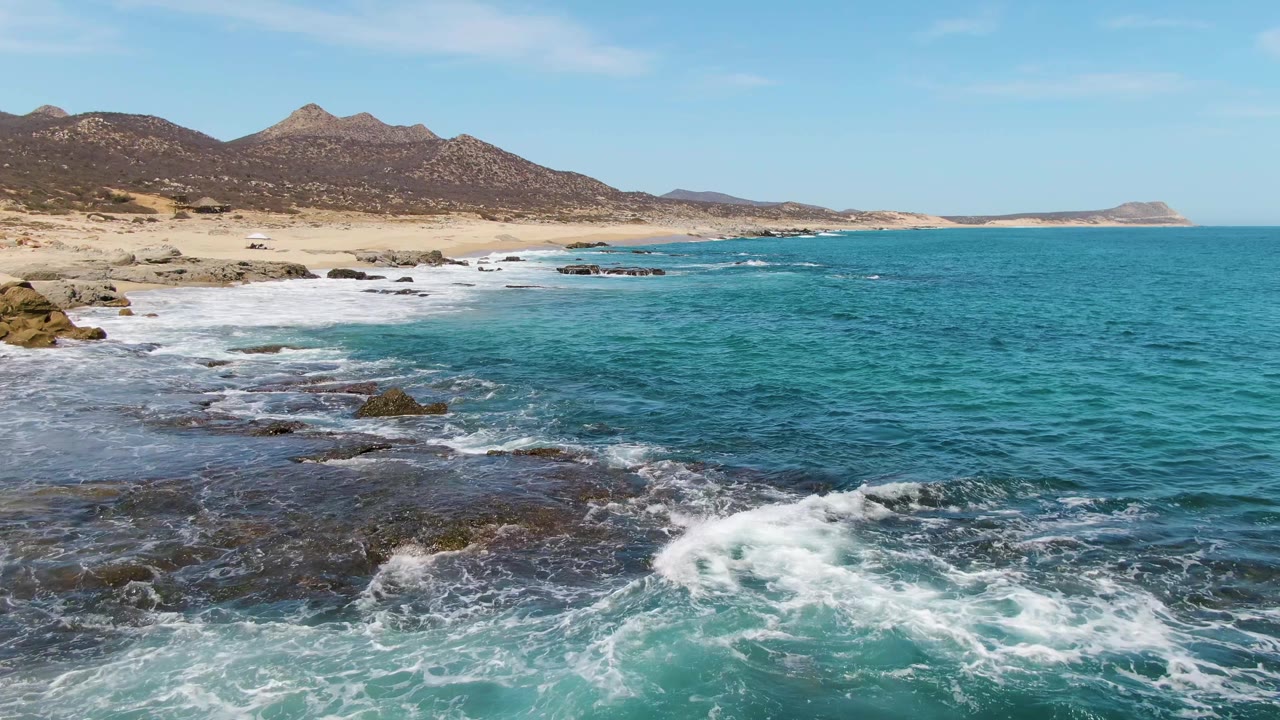 At 0:03, white water breaks across foreground rocks while the Buzzards Beach coastline and brown mountain ridge run across the background with no visible people.