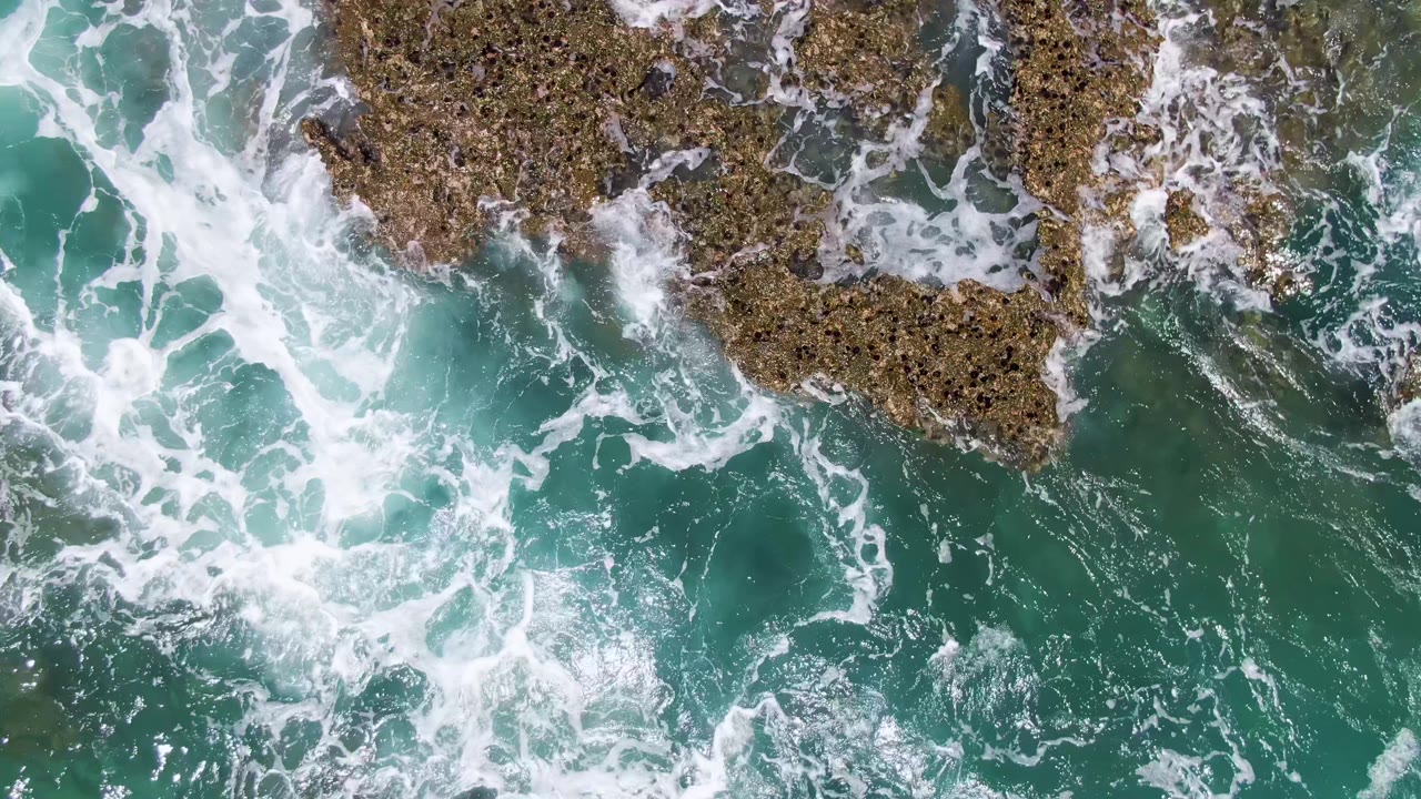 Overhead view at Buzzards Beach with dense white surf channels crossing green-blue water between several brown rock formations and no visible people.
