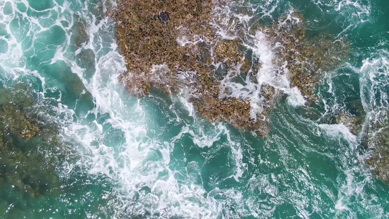 Top-down frame from Buzzards Beach showing churning turquoise water around darker submerged rock areas and exposed brown rock edges, with no visible people.