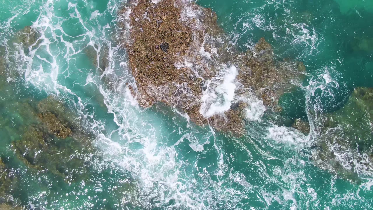 Overhead frame at Buzzards Beach where clear turquoise shallows reveal underwater rocks while white foam wraps around isolated brown outcrops, with no visible people.
