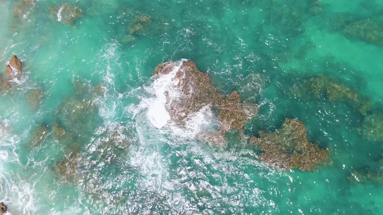 At Buzzards Beach, Baja California Sur, white foam spreads across the central rock while submerged darker rock shapes show through the turquoise water, with no visible people.